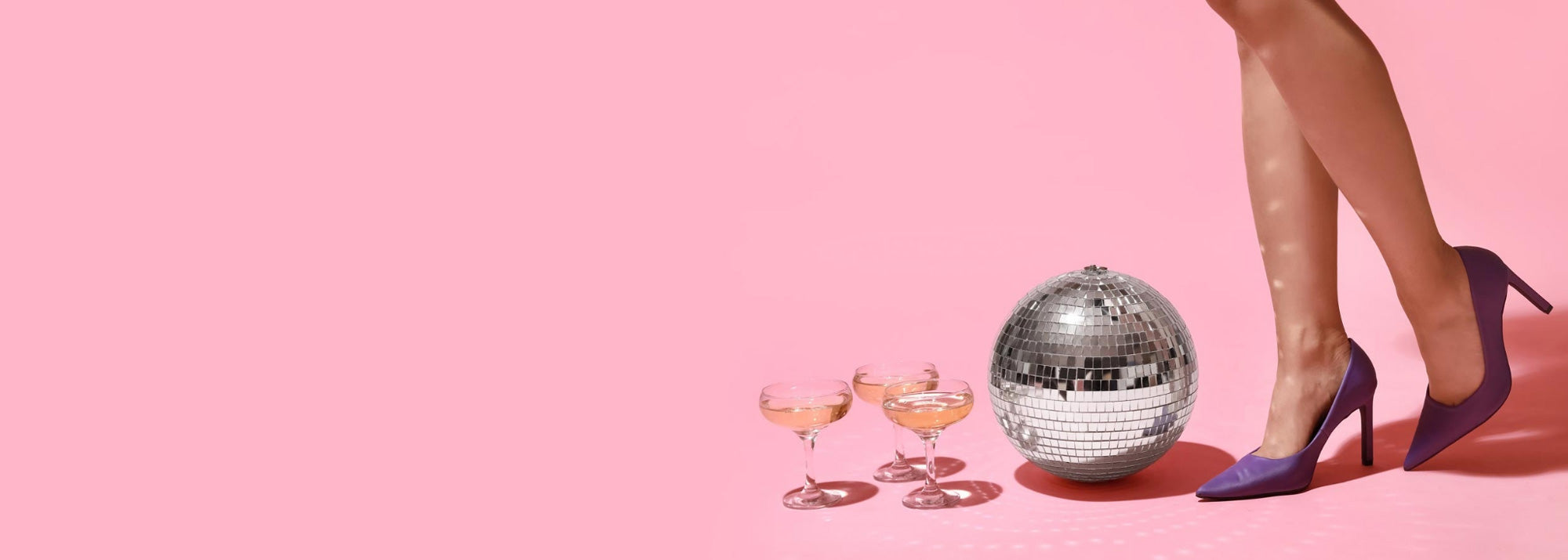 Tre champagneglas, en diskokula och kvinnoben i lila högklackade skor mot rosa bakgrund.